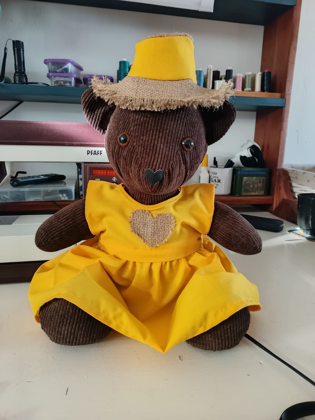 A brown corduroy teddy bear in a yellow cotton pinafore with a hessian heart on the front, wearing a hessian sun hat