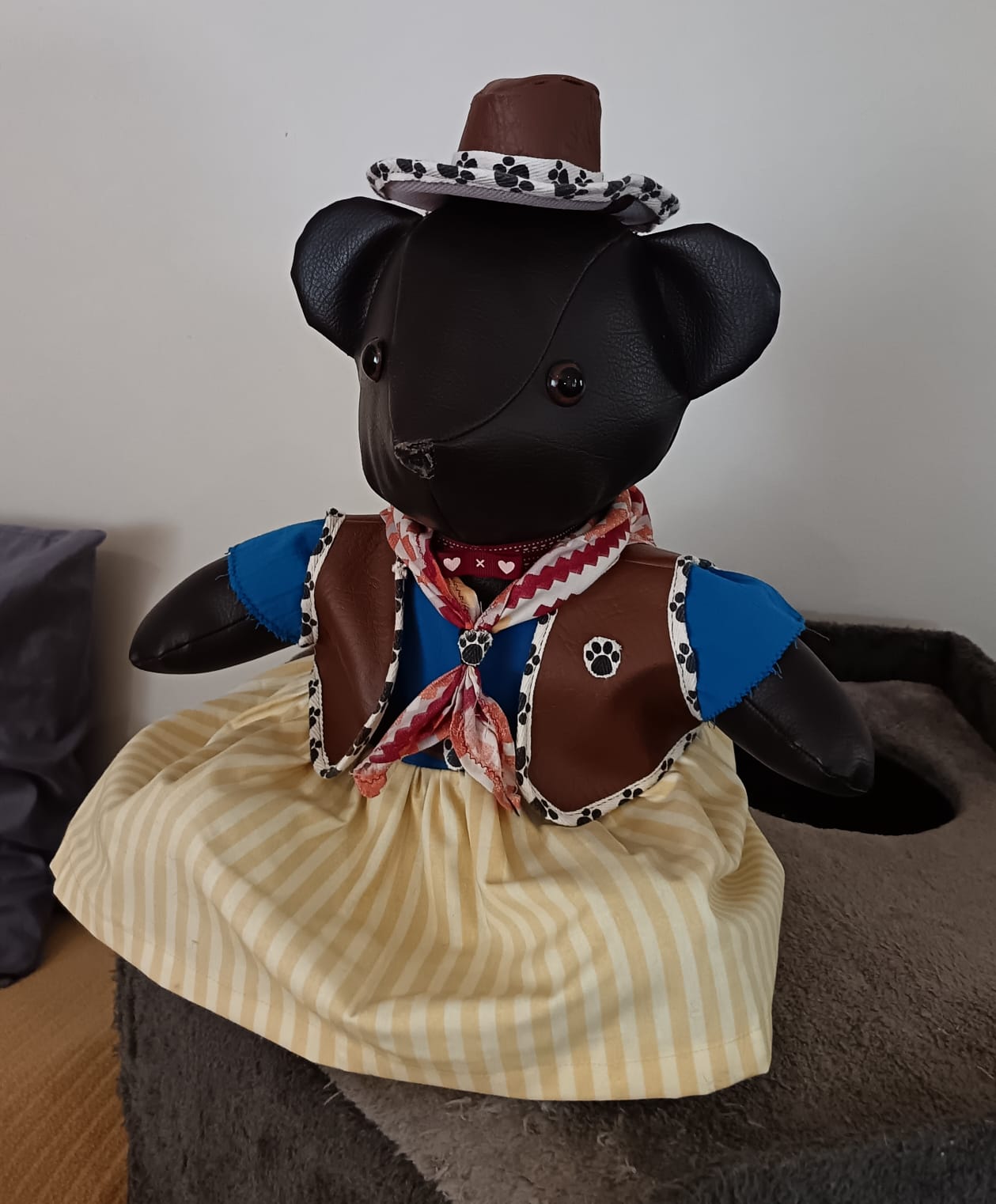 A dark brown leatherette teddy bear dressed as a cowgirl, in a brown waistcoat trimmed with paw-print ribbon, a yellow striped skirt, a neckerchief and a cowboy hat