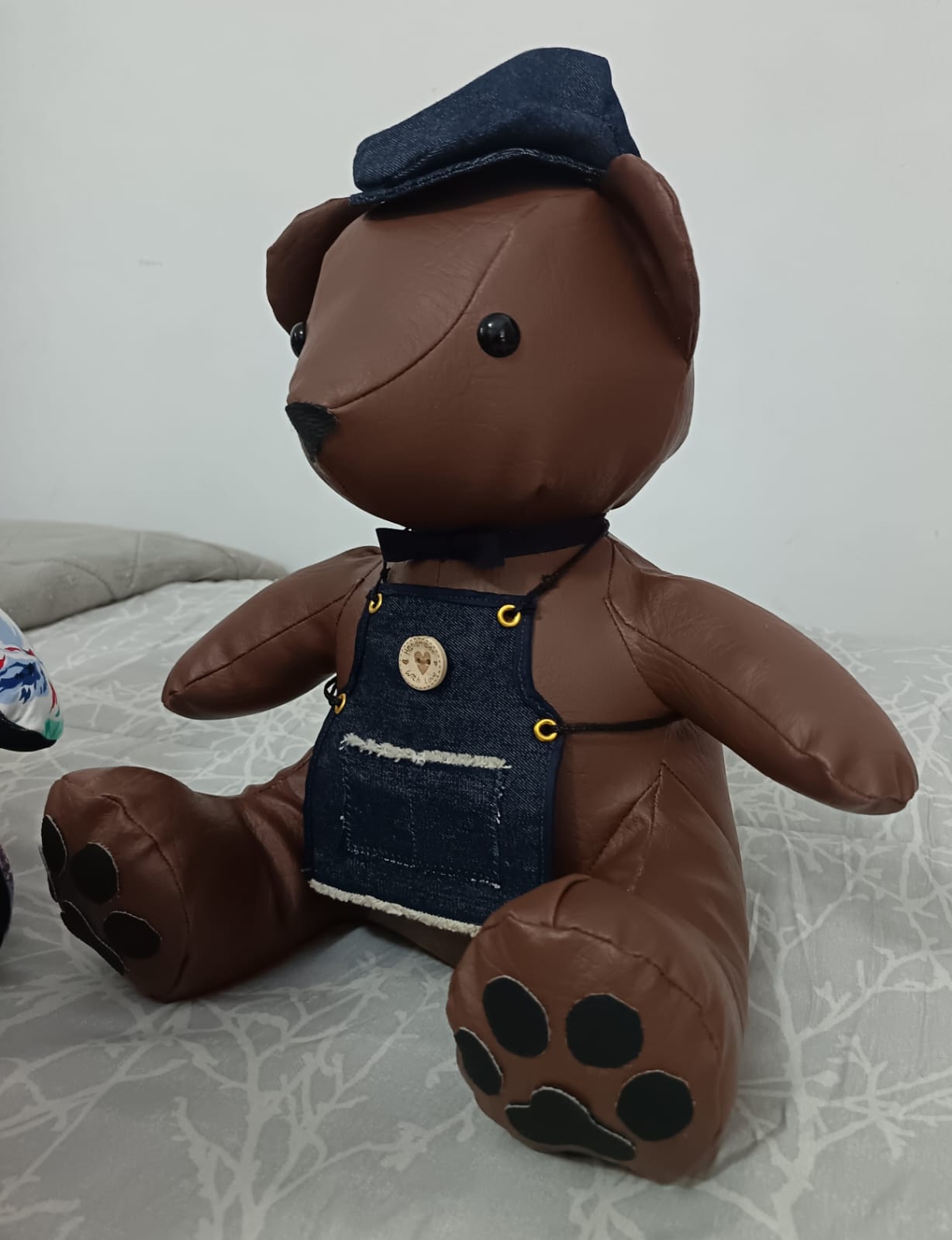 A brown leatherette teddy bear in a denim bib apron with brass eyelets, a frayed pocket and a wooden "handmade with love" button, wearing a denim flat cap and a navy bow tie