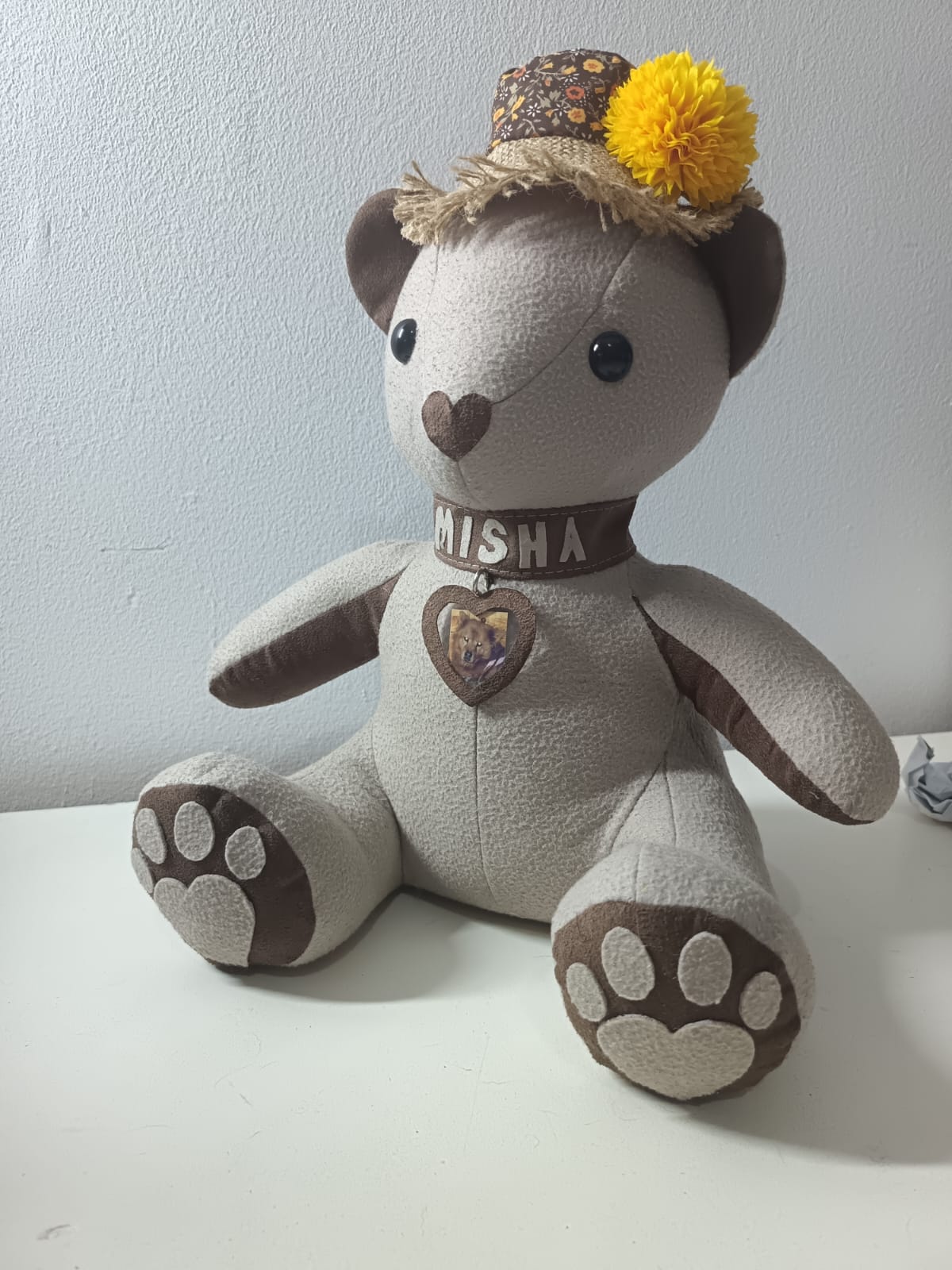 A cream and brown teddy bear with paw-print foot pads, a collar reading MISHA with a heart-shaped photo locket of a dog, and a straw hat with a yellow flower