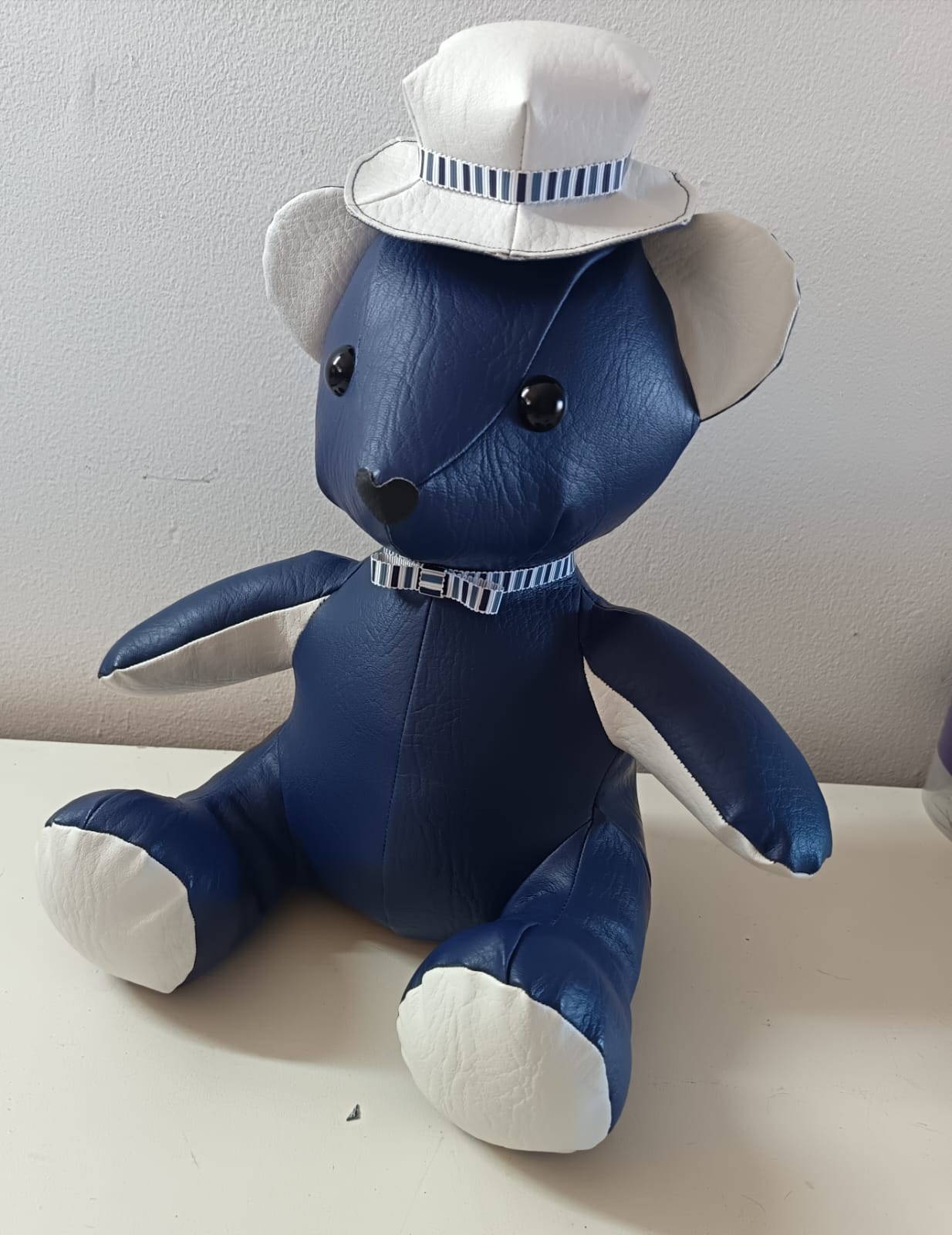 A navy leatherette teddy bear with white ears, inner arms and foot pads, wearing a white hat and a bow tie in matching blue and white striped ribbon
