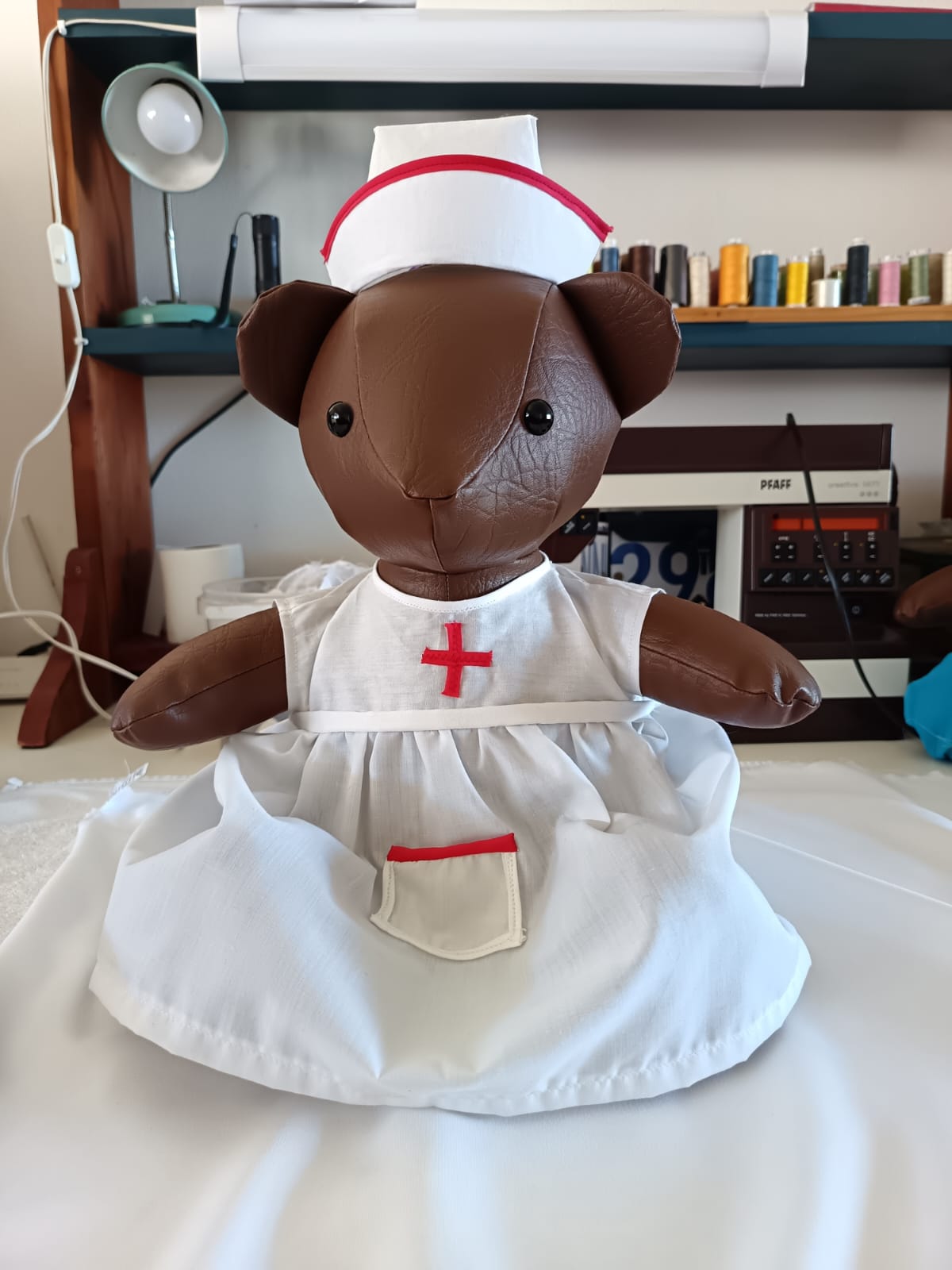 A brown teddy bear in a white nurse’s pinafore with a red cross, and a white cap with red trim