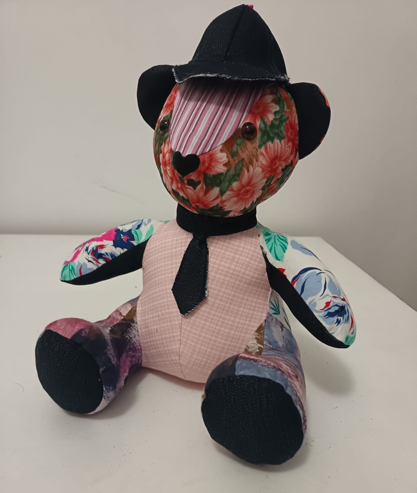 A patchwork teddy bear with a floral head, a striped muzzle, a pink check body, floral arms and painted-print legs, with black denim ears and foot pads, a black denim cap and a matching tie