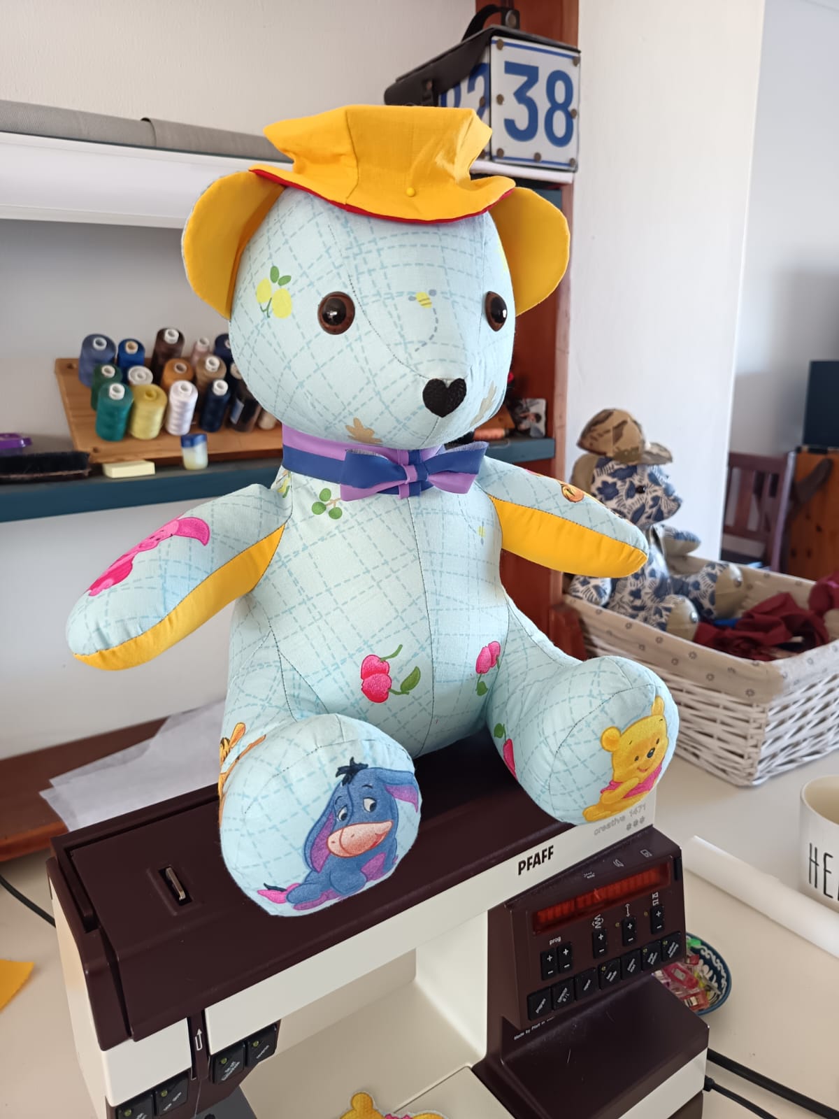 A pale blue teddy bear sewn from a nursery cot sheet printed with Winnie the Pooh characters, wearing a yellow sun hat and a ribbon bow