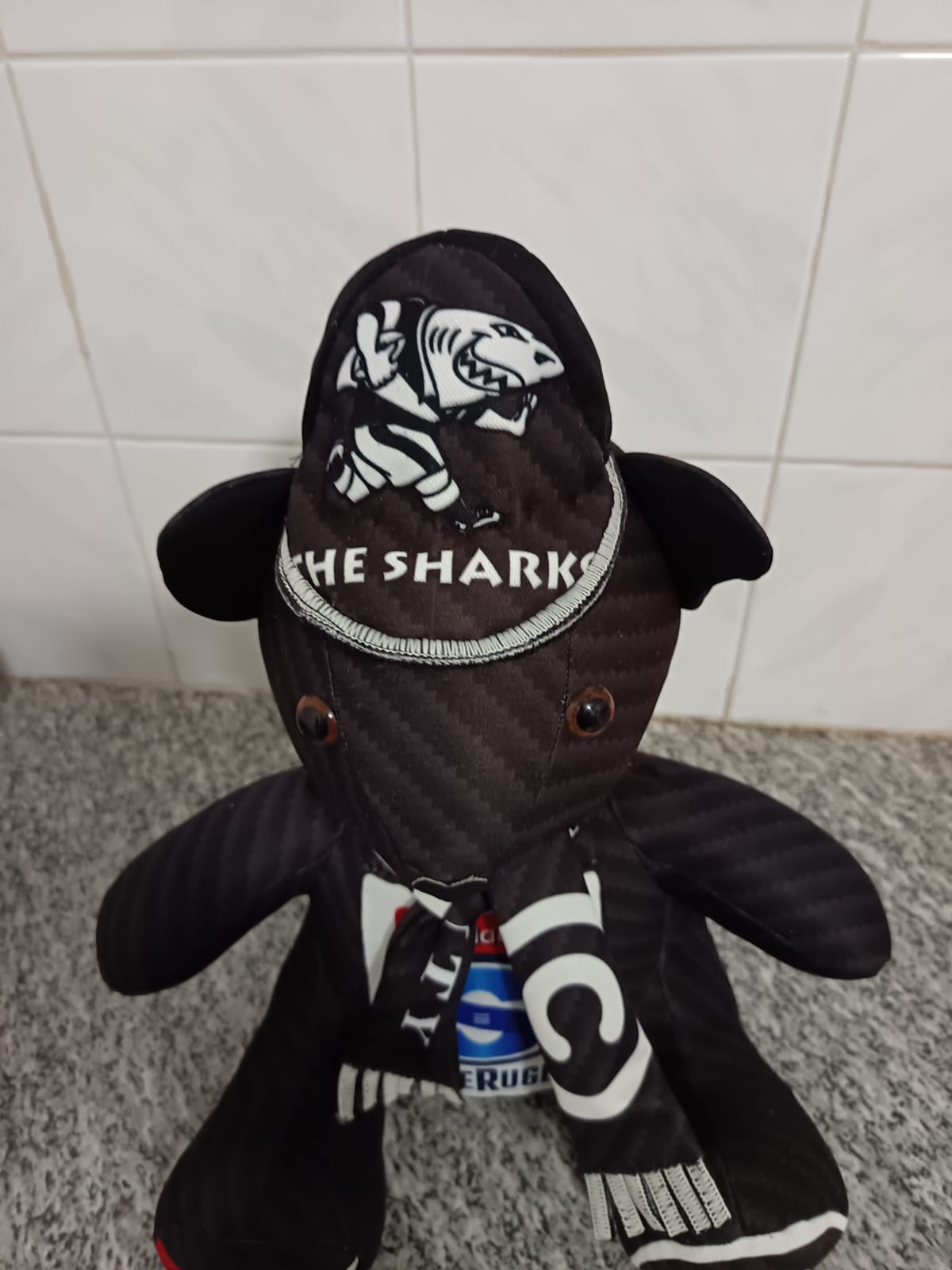 A black teddy bear sewn from a Sharks rugby jersey, wearing a hood with the Sharks emblem and a fringed team scarf