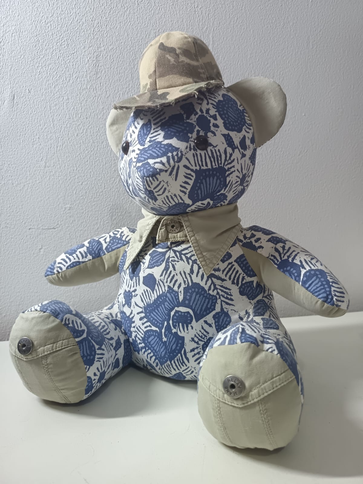 A teddy bear sewn from a blue and white printed shirt, with khaki ears, arms and foot pads, the khaki shirt collar at its neck, Relay Jeans buttons on its feet and a camouflage cap