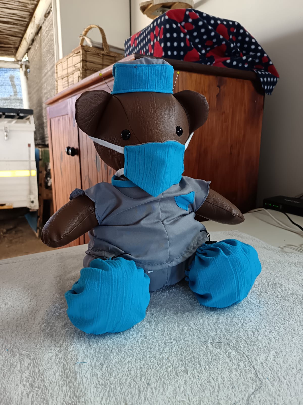 A brown teddy bear in grey theatre scrubs with a teal mask, scrub cap and booties, still pinned on the work table